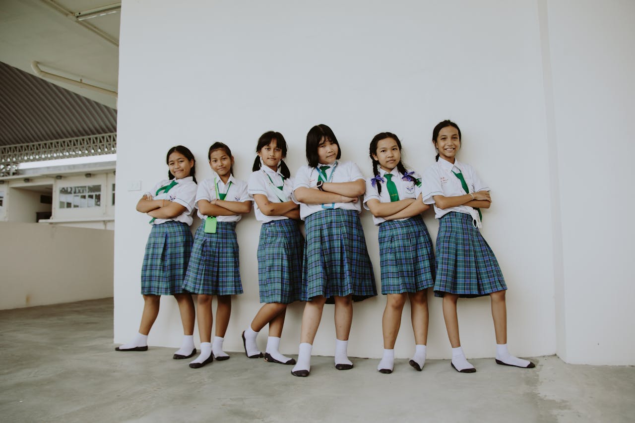 gallery-1 A group of six smiling schoolgirls in uniform standing indoors with arms crossed, displaying friendship.