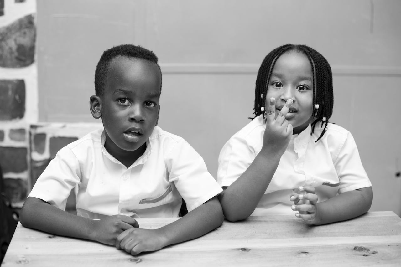 Monochrome image of two African children seated at a desk in school uniforms, sharing a candid moment.