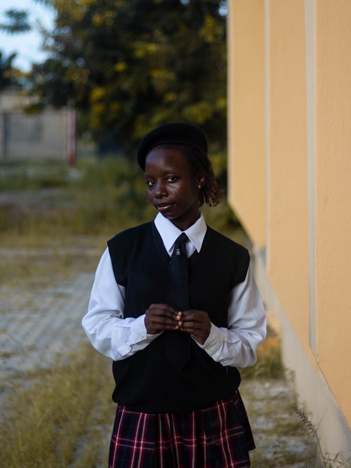 gallery-3 A young woman stands confidently in a school uniform next to a yellow building, displaying a thoughtful expression.