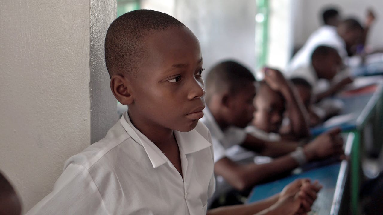 gallery-4 Portrait of a thoughtful young boy in a Haitian classroom, wearing a white shirt.